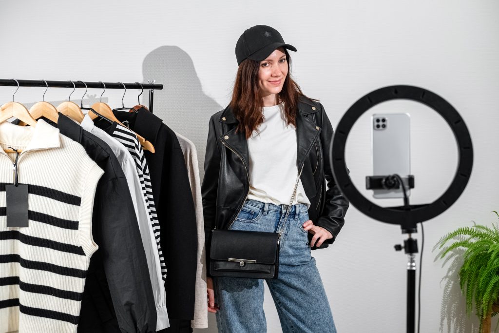 Fashion influencer creating social media content while modeling casual outfit with leather jacket, cap, and handbag in front of clothing rack and ring light.